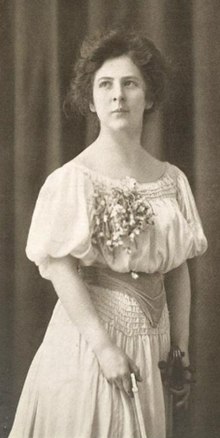 Eine junge weiße Frau in einem weißen Kleid, stehend; In ihren Händen sind Bogen und Hals einer Geige.