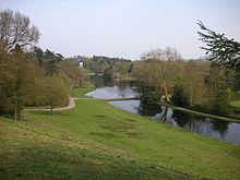 A long view from a hill with a lake stretching into the distance, beyond which rises another hill with a small white building on it. Trees and lawn border the lake.