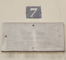 Fargebilde av en beige minneplate på en off-white vegg.  Gatenummer 7 er synlig øverst.