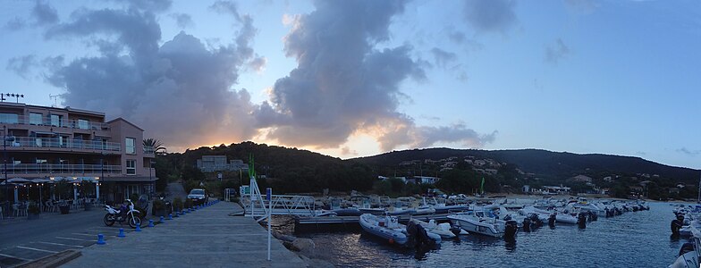 panorama montrant une petite marina avec des bateaux de petite taille et un hotel, en arriere plan des colines et des nuages éclairés par le soleil deja couché