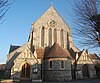 A low-roofed, wide, cobbled flint church with several sections, facing a road behind a wall of the same material. On the right, partly obscured, is an apse with plain stone-dressed lancets; next to it is the body of the church, with tall round-headed windows. In the foreground is a spirelet and a low extension with a five-light window.