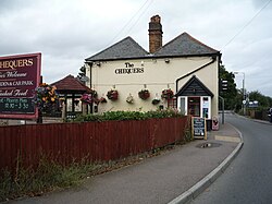 A photo of the Chequers Pub. It is a cream stucco two story wonky building on the bend in a road. A sign, saying "The Chequers" is displayed on the side of the building in a serif font.