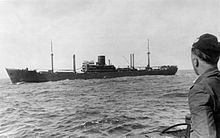 A merchant ship with a central superstructure and single funnel at sea. The photograph was taken from another vessel, with a sailor standing in frame on the right side, facing the merchant ship