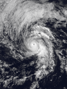 A well-organized hurricane over the Eastern Pacific Ocean, with spiral bands; a dense, white, round mass of clouds in the middle; and a clear eye at the center. A large mass of thinner clouds to the northwest of the hurricane curves in tandem with the hurricane's circulation.