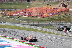 A matte navy single-seater open-wheel racing drives around a corner on a tarmac course while a grandstand full of people dressed in orange set off orange fireworks in the background.