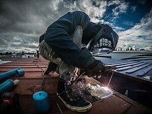 Schweißer mit Helm aber ohne spezielle Arbeitskleidung arbeitet auf einem Container. Im Hintergrund bewölkter Himmel