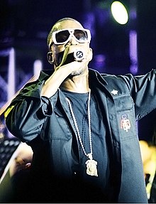 A man holding a microphone and wearing white sunglasses, black clothing and a chain around his neck.