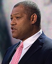 A man speaking in a black suit with a pink tie