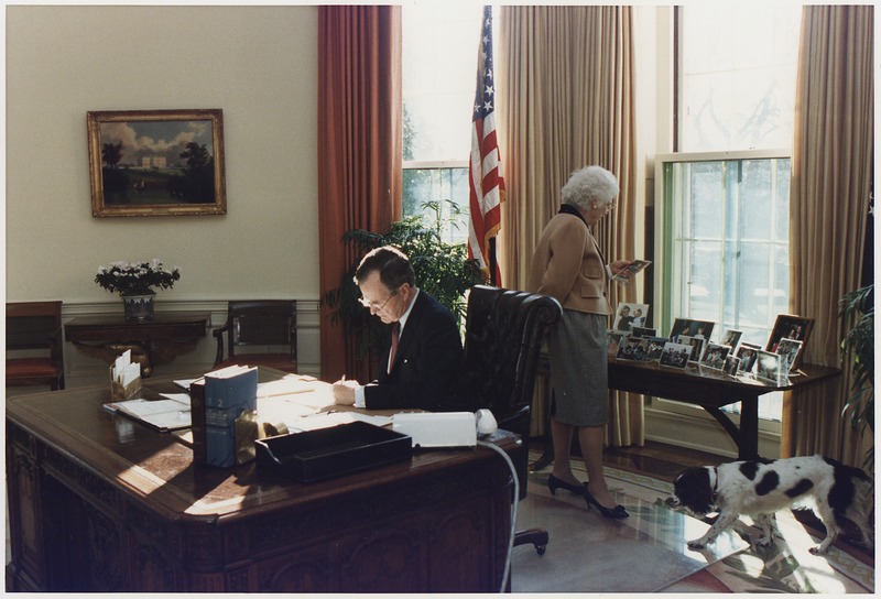 File:President Bush works at his desk in the Oval Office as Mrs. Bush looks at photographs on the table behind the Oval... - NARA - 186389.tif