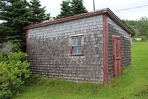 A small wooden building, with a slanting shed roof. One window is visible on one side, a Dutch door on the other, with a horseshoe nailed to the door. The building has red wooden trim around the corners, window and door, and the exterior walls are covered with grey wood shingles.