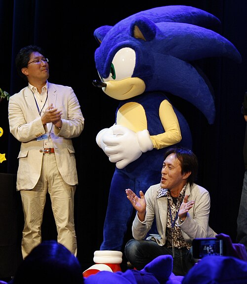 Sonic the Hedgehog co-creators: programmer Yuji Naka (left) and artist Naoto Ohshima (right) with a costume of the titular character (center)