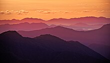 Une série de crêtes de montagnes, de noir, violet, un ciel orange, quelques rayons de soleil rasent les vallées.