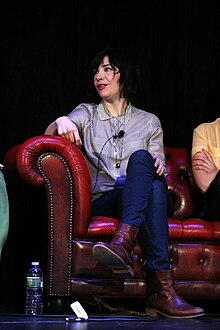 Carrie Brownstein sits on a leather couch with her right arm on the armrest and her legs crossed at the knee. She is speaking to someone who is outside the frame. She wears boots, dark jeans, and a blouse with thin vertical stripes. A lavalier microphone is clipped to her blouse.