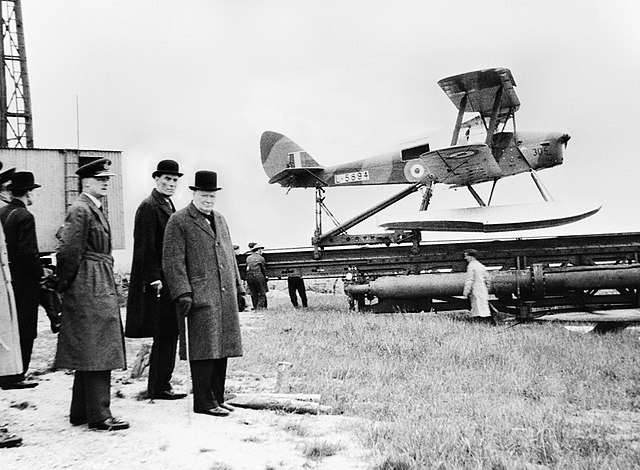 Winston Churchill, David Margesson, and others waiting to watch the launch of a DH.82 Queen Bee target drone, 6 June 1941