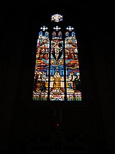Photographie couleur d'un vitrail d'église montrant un Christ crucifié entouré d'anges sous lequel une eucharistie est célébrée.