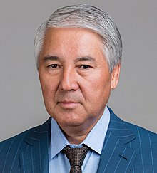 An older man of Central Asian complexion with grey hair, looking seriously into the camera with a grey background.