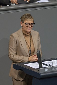 Sabine Dittmar steht an einem Pult und trägt kurzes blondes Haar. Ihr Anzug ist beigefarben und sie trägt eine Brille.