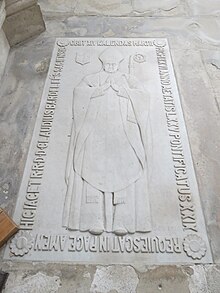 Photo de longue dalle sculptée en bas relief représentant un évêque mitré, avec inscription funéraire sur le pourtour.