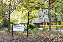 Eingang zum Berufsförderungswerk Düren, ein Schild mit Firmen-Beschriftung und Logo steht unter einem Baum im Vorgarten zum BFW