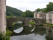 Fotografía en color de un puente de dos arcos que se refleja en un río.  Está ubicado en un pueblo verde.