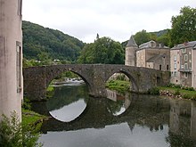 Foto a colori di un ponte a due arcate che riflette in un fiume.  Si trova in un villaggio verde.