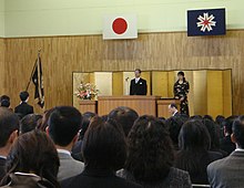 A group of people facing a man and woman on a stage. Two flags are above the stage.