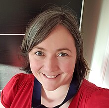 Photograph of Dr Kirsty Ross from the shoulders up. She is a white woman smiling at the camera whilst wearing a red dress.