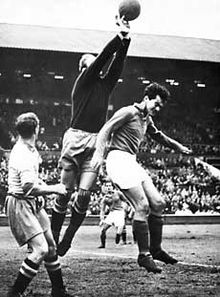 Dans une photo en noir et blanc, le gardien s'envole dans les airs, pour attraper le ballon, au détriment d'un joueur adverse.