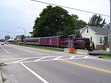 A train parked along a railroad track, in front of and parallel to a road. The train includes a purple and black diesel locomotive numbered 6, a baggage car, and several passenger cars labeled "Newport Dinner Train".