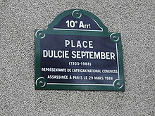 Imagem da placa toponímica da Praça Dulcie September, no no 10º Arrondissement de Paris. A placa tem uma forma rectangular em baixo, com uma meia lua por cima, com uma moldura verde e branca, e letras bramcas sobre um fundo azul. O texto na placa inclui a referência ao arrondissement, o nome da praça, as datas de nascimento e morte de Dulcie (1935-1988) e a descrição Representante do Congresso Nacional Africano assassinada em Paris a 29 de março de 1988.