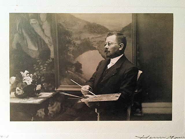Jules-Alexis Muenier, der Maler in seinem Atelier. Sitzender Mann mit Schnurrbart und Brille mit der Malerpalette in der Hand vor einem Landschaftsgemälde.