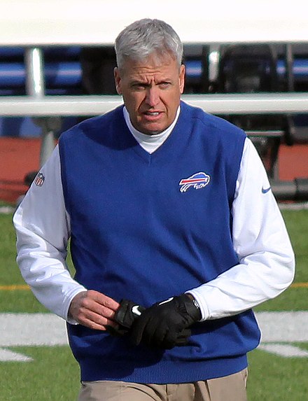 Color photograph of Rex Ryan on football field, wearing a royal blue v-neck sweater vest with a Buffalo Bills logo, over a white long-sleeved t-shirt.
