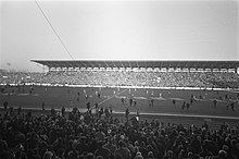 Photographie en noir et blanc. Vue élevée des tribunes d'un stade au cours d'un match.