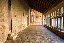 Photo couleur d'une galerie de cloitre entre un mur de briques à gauche et une colonnade à arc roman à droite, donnant sur le jardin. Le sol est pavé de galets et la toiture simple penche vers le jardin.
