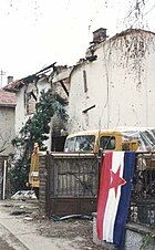 A blue-white-red Yugoslav flag, with the red Communist star in the middle, hangs on an iron fence outside the ruined shell of a house. A truck is partly visible parked in the driveway next to the building.