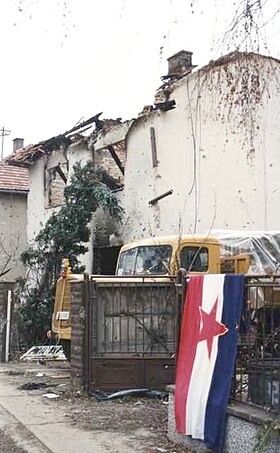 청백적에 가운데 공산주의를 뜻하는 별이 그려진 유고슬라비아의 국기가 포격을 받아 파괴된 집 바깥 철조망 담에 걸린 모습. 건물 철담 뒷편에 트럭이 주차되어 있는 모습이 보인다.