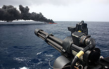 Photograph of a drug vessel burning in the background, with a ship-mounted minigun in the foreground.
