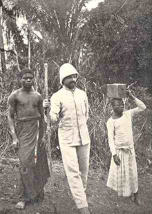 Émile Laurent dans la nature au Congo, vêtu de blanc, avec un casque colonial, au centre entre deux Congolais en tenues traditionnelles.