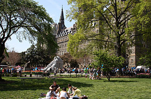 Many students mingle in the background while a group sit in the foreground on a grass lawn. The large stone clocktower is seen above the trees on the lawn.