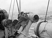 A lookout of a convoy escort, posing his binoculars on a depth charge thrower with which depth charges were launched to the sides of the escort HMS Viscount depth charge thrower 1942 IWM A 13362.jpg