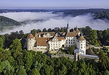 Vue aérienne d'un château à la toiture rouge niché dans une forêt, avec de la brume dans le fond de l'image.