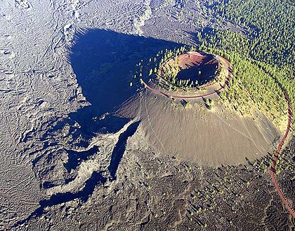 Aerial photograph of a cinder cone butte casting a shadow over a large lava field. A road enters from the side of the photo and spirals to the top of the butte.