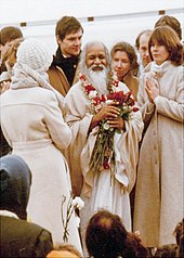 A photograph of the Maharishi holding flowers and surrounded by people wearing coats.