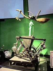 Un balai avec un siège et des cales pour les pieds est positionné au sommet d'une machine à pompes, davant un fond vert.