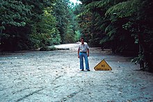 A high surface of mud with a person posing next to a traffic sign reading "NO SHOULDER" that is almost completely buried.