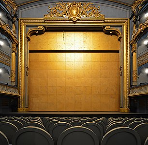Un grand rideau doré ferme la scène. On distingue les fauteuils du parterre et les balcons.
