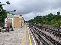 A man with black hair, a black shirt, black pants, and black shoes reading a newspaper and sitting on a brown bench on a grey railway platform