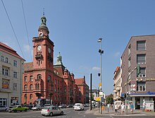 Farbfotografie mit Blick auf eine vierspurige Straße, die zu einem großen roten Gebäude mit auffälligem Uhrenturm führt.