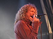 Um homem com cabelo comprido e cacheado, vestindo uma camisa vermelha e cantando em um microfone em um pedestal.