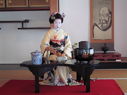 A maiko does a ryūrei style tea where a table and chair are used; visible from left to right are the fresh water container, caddy, bowl, and iron pot.